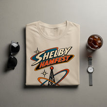 Load image into Gallery viewer, Light tan t-shirt with "Shelby Hamfest" logo featuring a radio tower and orbiting rings.