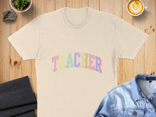 Load image into Gallery viewer, Cream t-shirt with pastel rainbow "TEACHER" text, flat lay with coffee and denim jacket.