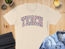 Load image into Gallery viewer, Cream t-shirt with "TEACH" in floral letters, displayed on a wooden surface.