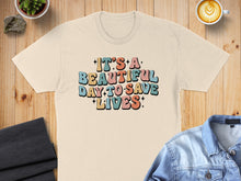 Load image into Gallery viewer, Cream t-shirt with colorful text "It's a beautiful day to save lives".
