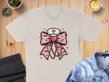 Load image into Gallery viewer, Pink ribbon with stethoscope, heart, and nurse cap graphic on a tan t-shirt.
