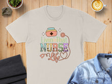 Load image into Gallery viewer, "Livin' That Nurse Life" text with nurse cap and stethoscope on a light t-shirt.