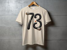 Load image into Gallery viewer, Cream t-shirt with a black hose forming the number 73 and a red dot.