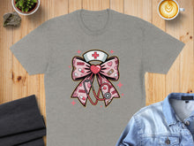 Load image into Gallery viewer, Grey t-shirt with a pink bow graphic featuring a cross, stethoscope, and bandages.