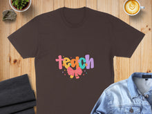 Load image into Gallery viewer, Brown t-shirt with colorful "teach" text and a pink bow graphic.