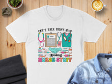 Load image into Gallery viewer, "CAN'T TALK RIGHT NOW DOING NURSE STUFF" text with medical icons on a white t-shirt.