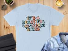 Load image into Gallery viewer, Light blue t-shirt with colorful "It's a beautiful day to save lives" text.