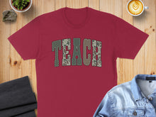 Load image into Gallery viewer, Maroon t-shirt with "TEACH" in camouflage lettering, laid flat with accessories.