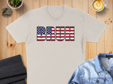 Load image into Gallery viewer, Tan t-shirt with "BRUH" in American flag design on a wooden surface.