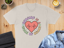 Load image into Gallery viewer, Cream t-shirt with "Nursing is a work of heart" text and a heart monitor graphic.