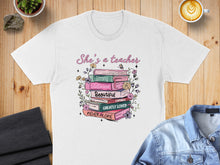 Load image into Gallery viewer, White t-shirt with "She's a teacher" and uplifting words on stacked books.