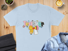 Load image into Gallery viewer, Light blue t-shirt with colorful "Teacher" text, pencil, and school-themed graphics.