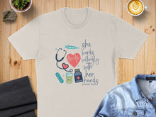 Load image into Gallery viewer, Cream t-shirt with stethoscope, heart, syringe, and "she works willingly with her hands" text.