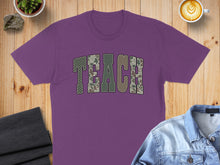 Load image into Gallery viewer, Purple t-shirt with "TEACH" in camouflage letters, laid flat with coffee and jeans.
