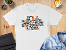 Load image into Gallery viewer, White t-shirt with colorful retro text reading "It's a beautiful day to save lives."
