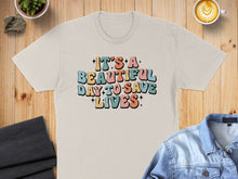 Load image into Gallery viewer, T-shirt with colorful retro text "It's a beautiful day to save lives".
