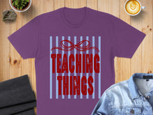 Load image into Gallery viewer, Purple t-shirt with red text "TEACHING THINGS" on blue stripes.
