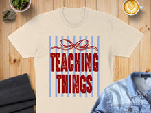 Load image into Gallery viewer, Cream t-shirt with "TEACHING THINGS" in red font and blue stripes.
