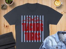 Load image into Gallery viewer, Dark gray t-shirt with "TEACHING THINGS" in distressed red font on a striped background.

