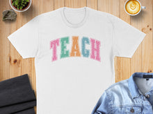 Load image into Gallery viewer, White t-shirt with colorful "TEACH" text, laid flat with coffee and denim jacket.
