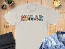 Load image into Gallery viewer, Cream t-shirt with colorful "NURSE" text, laid flat with coffee and denim jacket.
