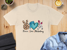 Load image into Gallery viewer, Cream t-shirt with "Peace Love Phlebotomy" text and heart, peace sign, and syringes graphic.
