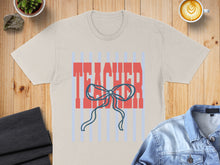 Load image into Gallery viewer, Cream t-shirt with "TEACHER" text and blue bow, on a wooden surface.
