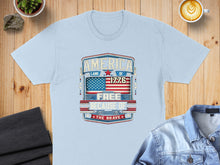 Load image into Gallery viewer, Light blue t-shirt with "America Land of the Free Because of the Brave 1776" graphic.
