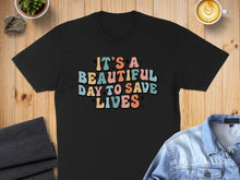Load image into Gallery viewer, Black t-shirt with colorful retro text: "IT'S A BEAUTIFUL DAY TO SAVE LIVES".
