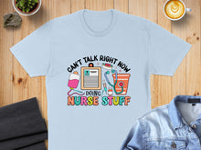 Load image into Gallery viewer, Light blue t-shirt with "Can't Talk Right Now Doing Nurse Stuff" text and medical icons.
