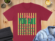 Load image into Gallery viewer, Burgundy t-shirt with green text "YOU ARE" and inspirational words, on a wooden surface.
