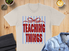 Load image into Gallery viewer, Cream t-shirt with red text "TEACHING THINGS" and a red bow design.

