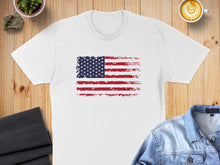 Load image into Gallery viewer, White t-shirt with distressed American flag graphic, laid flat on a wooden surface.
