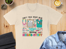 Load image into Gallery viewer, Cream t-shirt with "Can't talk right now, doing nurse stuff" and medical icons.

