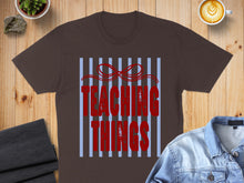Load image into Gallery viewer, Brown t-shirt with red "Teaching Things" text on a blue striped background.

