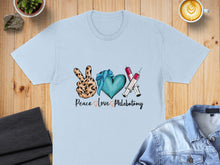 Load image into Gallery viewer, Light blue t-shirt with "Peace Love Phlebotomy" text and medical icons.
