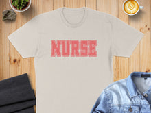 Load image into Gallery viewer, Cream t-shirt with "NURSE" in coral letters, laid flat with coffee and denim jacket.
